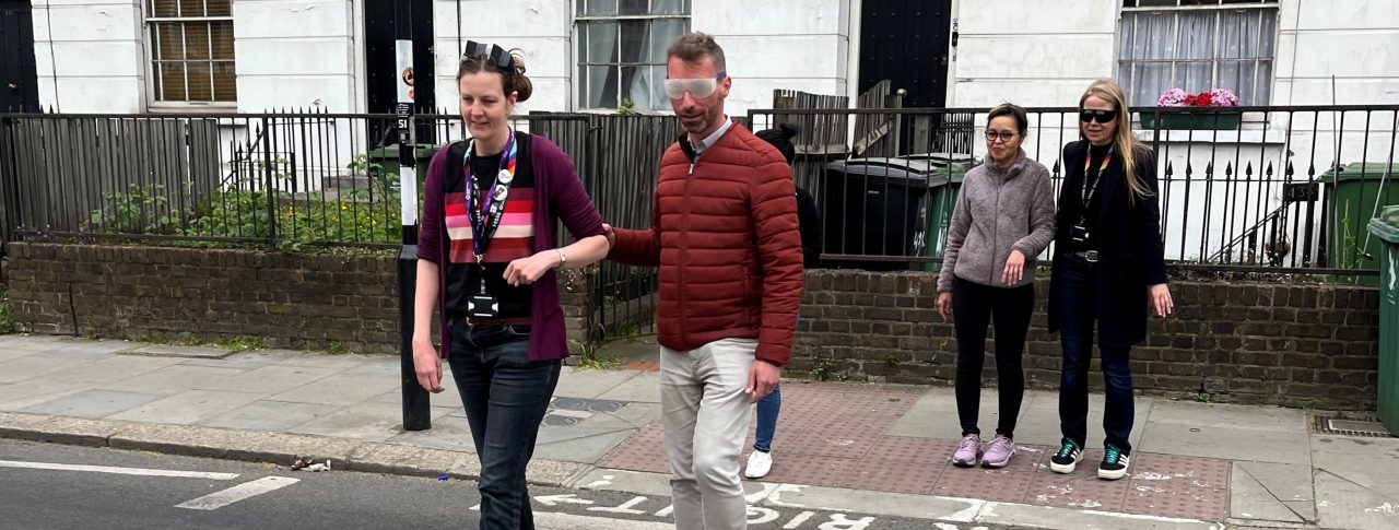 Camden Council councillors and cabinet members crossing a road at a zebra crossing during the sim spec walk. From left to right: Jenny Mulholland Camden Councillor (Gospel Oak ward), Adam Harrison, Cabinet member for a sustainable Camden and Camden Councillor (Bloomsbury ward), a My Sighted Guide Volunteer, and Sian Berry, Camden Councillor (Highgate Ward)