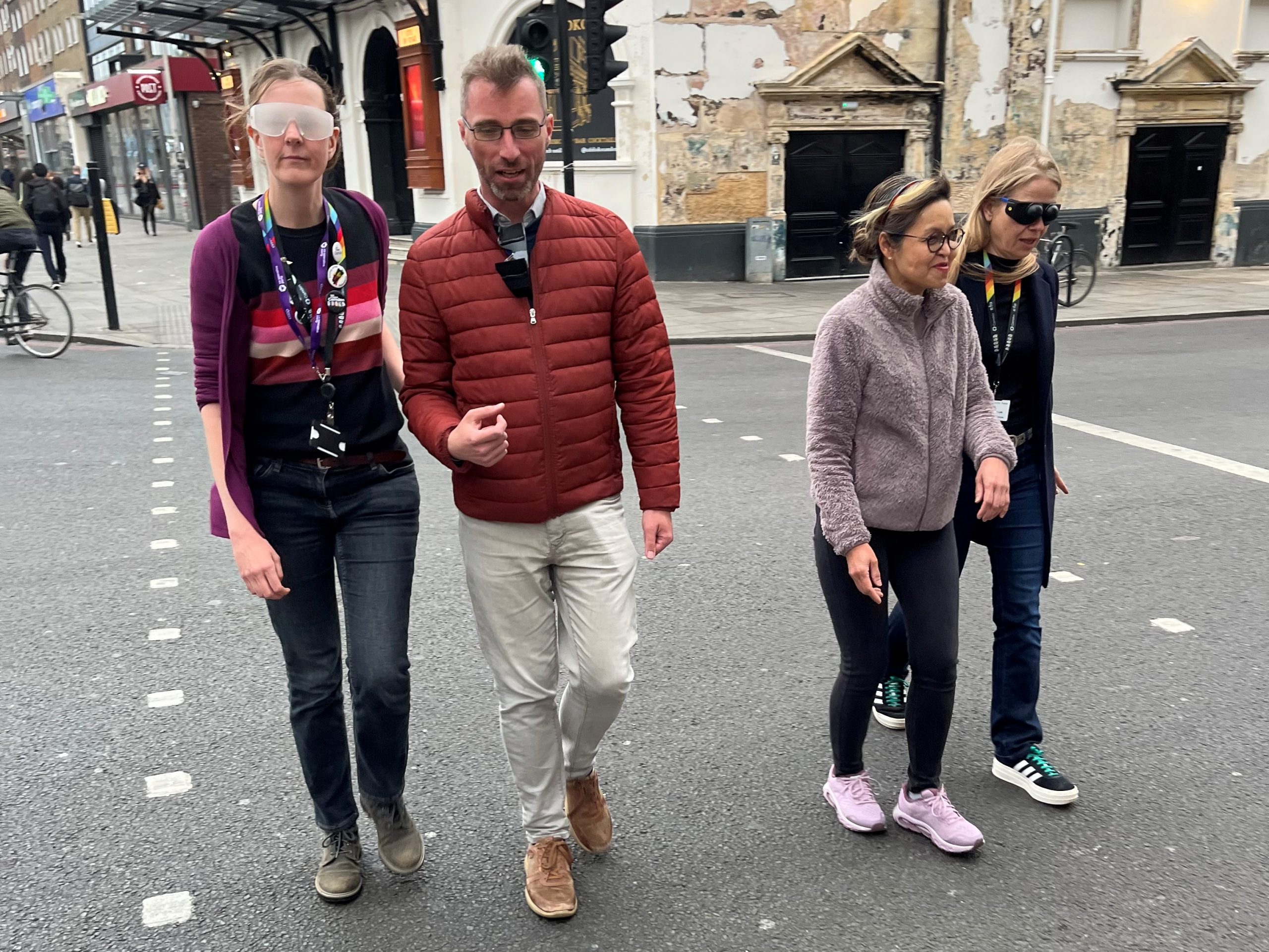 Camden Council councillors and cabinet members crossing a road during the sim spec walk at a pedestrian crossing. From left to right: Jenny Mulholland Camden Councillor (Gospel Oak ward), Adam Harrison, Cabinet member for a sustainable Camden and Camden Councillor (Bloomsbury ward), a My Sighted Guide Volunteer, and Sian Berry, Camden Councillor (Highgate Ward)