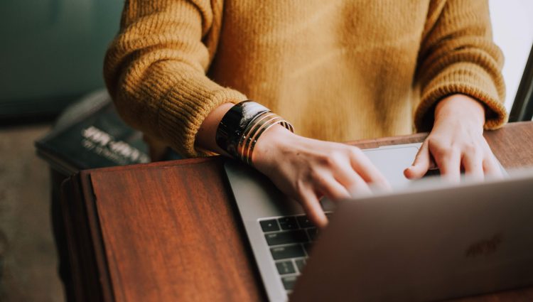 Close up of a woman's hands, typing on a silver laptop. She is wearing a mustard yellow jumper, and has two bangles on her wrist.