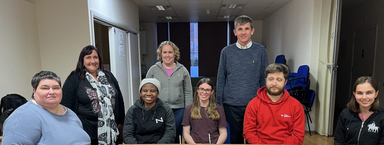 East Sussex SLC members at their 1st birthday meeting. They are sat around a table, with two birthday cakes on it. From left to right: Iris Keppler, Elaine Dampier, Ola Adeleke, Linn Davies, Lauren Eade, Dave Smith, Rich Wheeler, and Zehra Yunel.