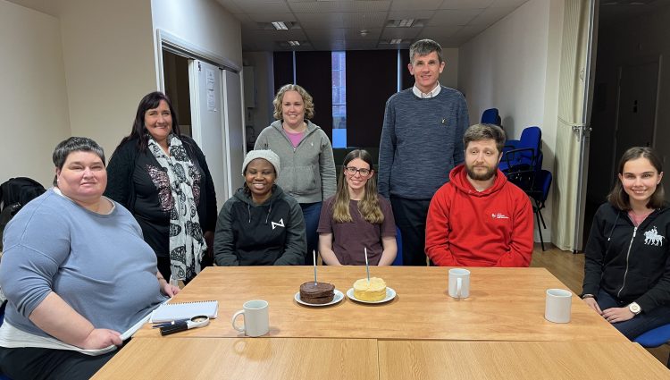 East Sussex SLC members at their 1st birthday meeting. They are sat around a table, with two birthday cakes on it. From left to right: Iris Keppler, Elaine Dampier, Ola Adeleke, Linn Davies, Lauren Eade, Dave Smith, Rich Wheeler, and Zehra Yunel.