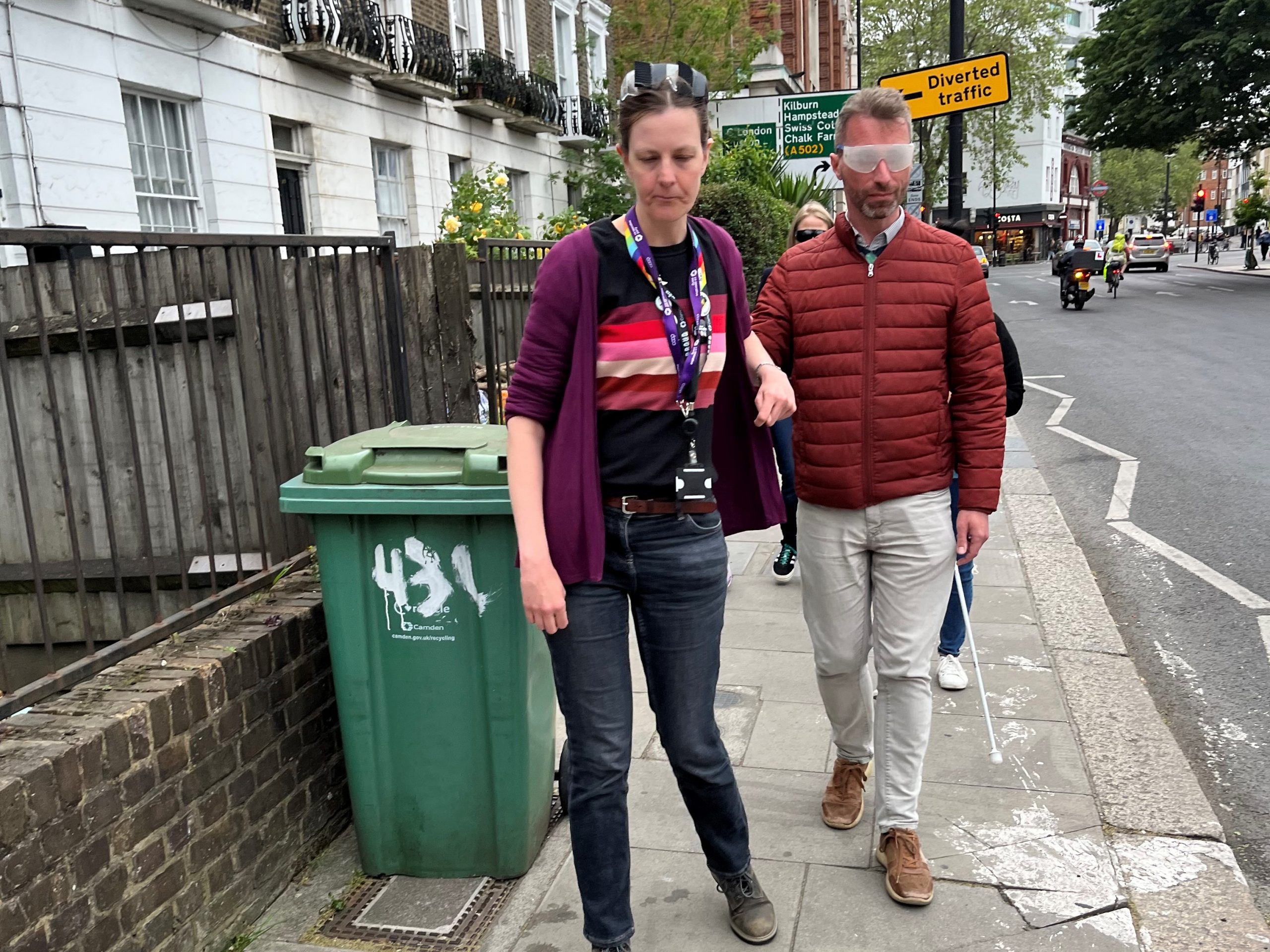 Jenny Mulholland, Camden Councillor (Gospel Oak ward), is guiding Adam Harrison, Cabinet member for a sustainable Camden and Camden Councillor (Bloomsbury ward) during the sim spec walk. She is guiding him around a green recycling bin which is sitting on the pavement.