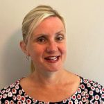 headshot of Kelly Barton, Engagement Manager for the north west. Kelly is standing against a white wall and smiling at the camera. Her hair is pulled back into a pony tail. She is wearing a black top with red and white flowers on it.