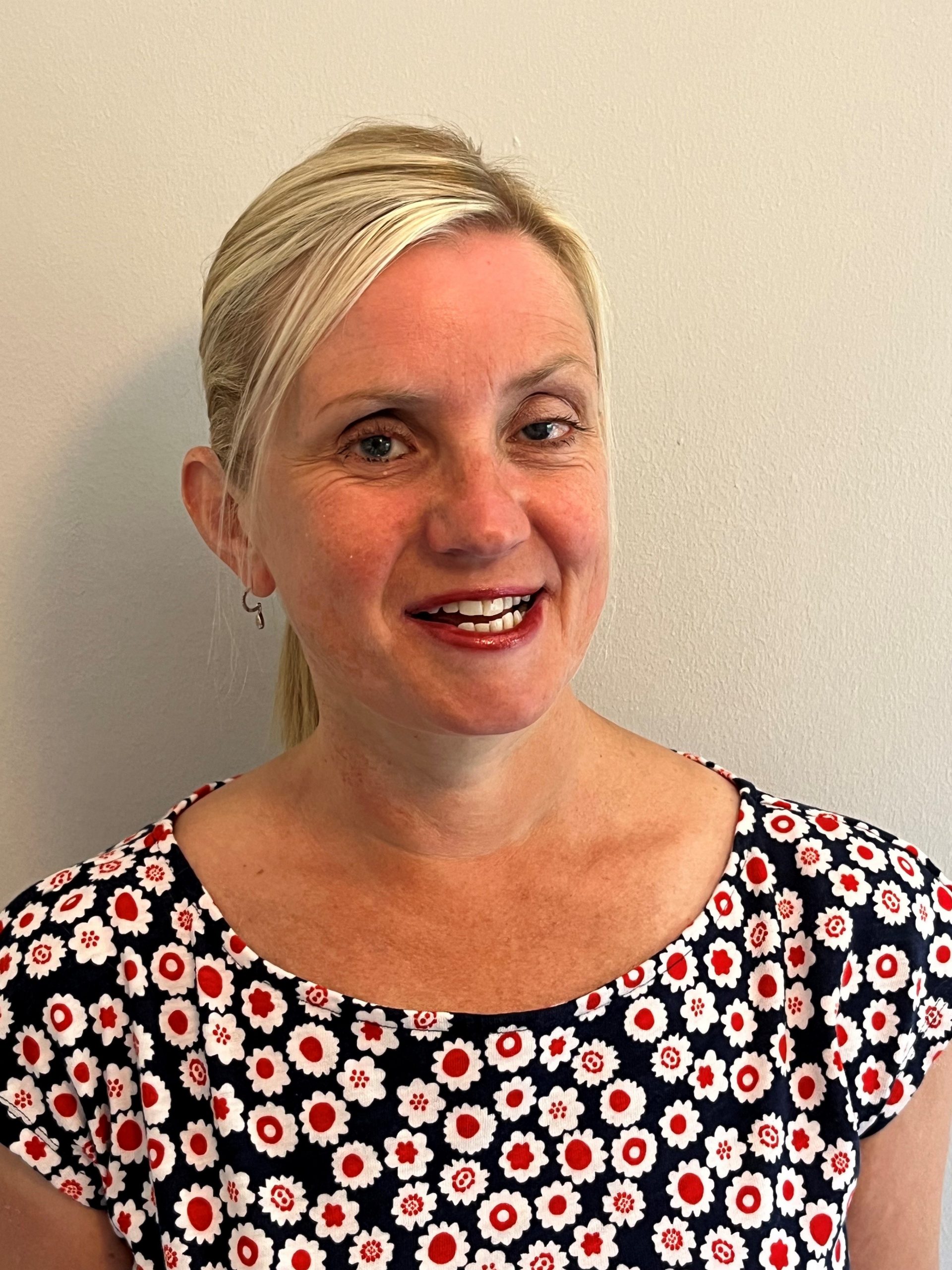 headshot of Kelly Barton, Engagement Manager for the north west. Kelly is standing against a white wall and smiling at the camera. Her hair is pulled back into a pony tail. She is wearing a black top with red and white flowers on it.