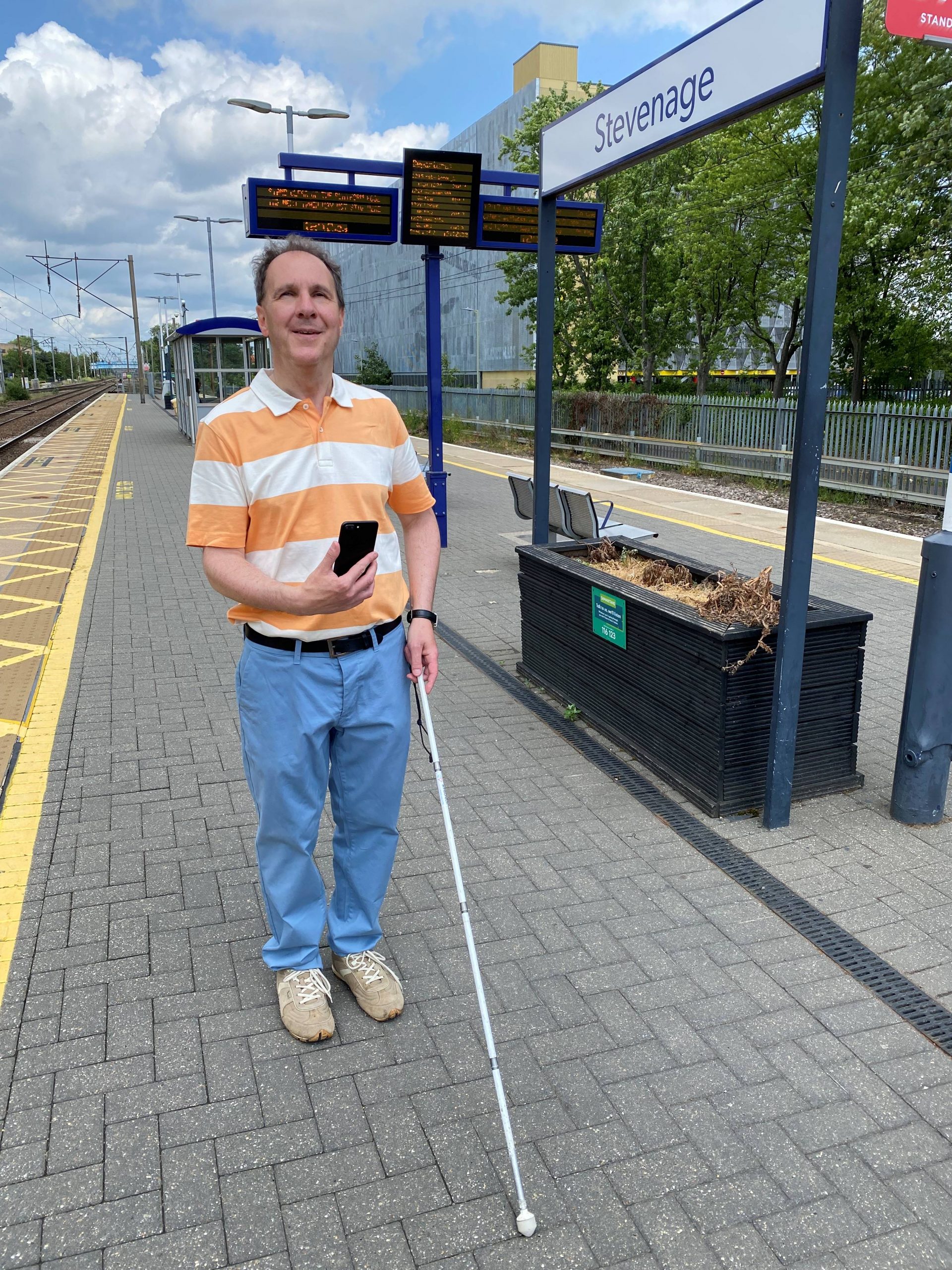 Paul Day, Bedfordshire SLC member, is standing on a platform at Stevenage train station. he is holding his long cane in one hand, and his phone up in the other.