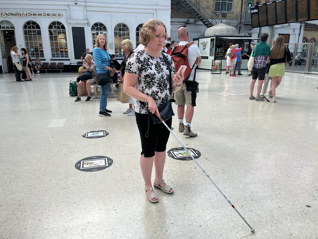 East Sussex SLC member, Linn Davies, standing in Brighton Train Station during our trials with Govia Thameslink. She is holding her cane in one hand, her phone in the other, using a wayfinding app.