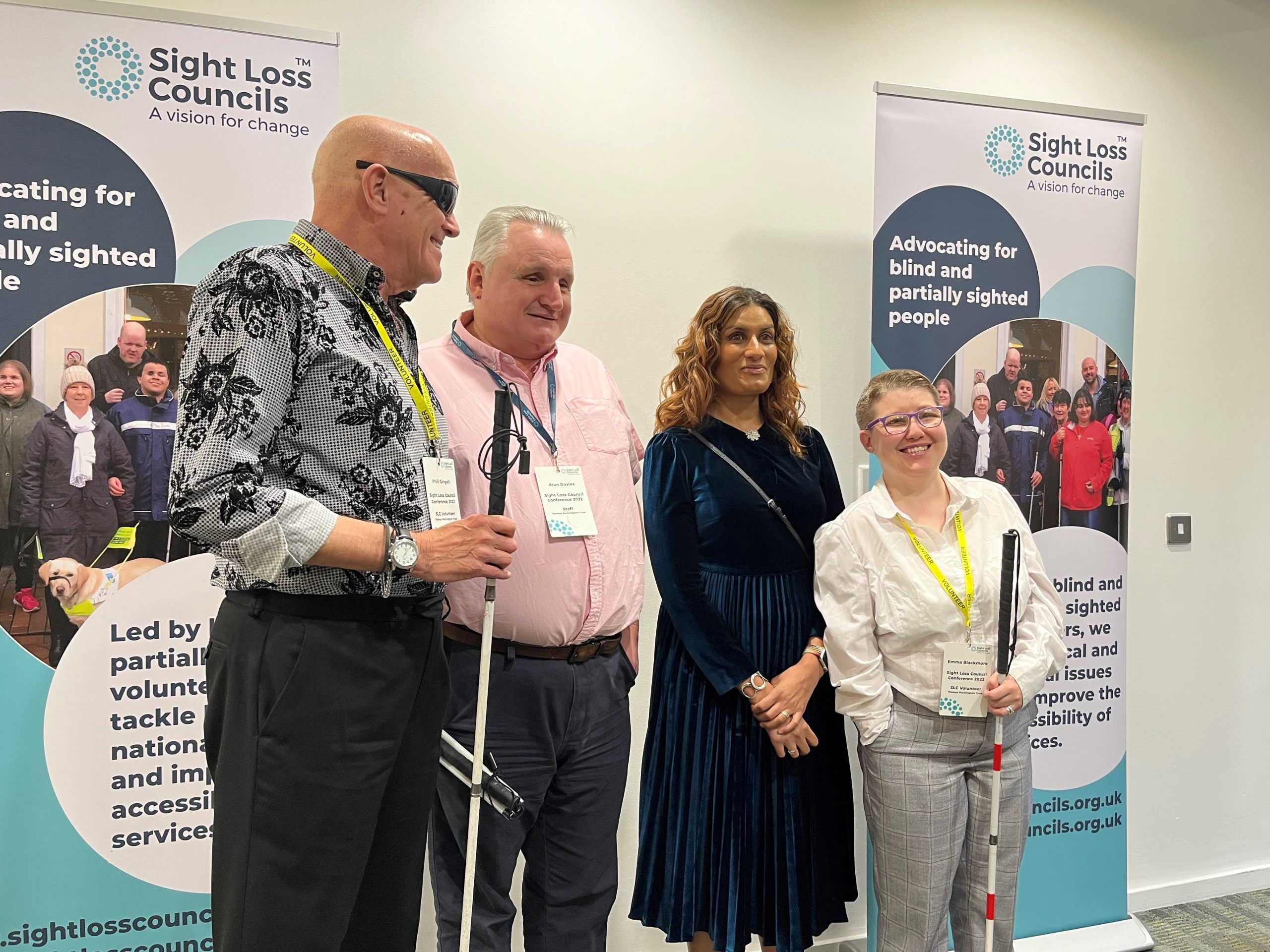 From left to right: West of England SLC member, Phil, Engagement Manager Alun Davies, and SLC members Anela and Emma. They are stood together at the 2022 Sight Loss Council conference in front of the SLC banners. They are having their photo taken by the official photograpaher.
