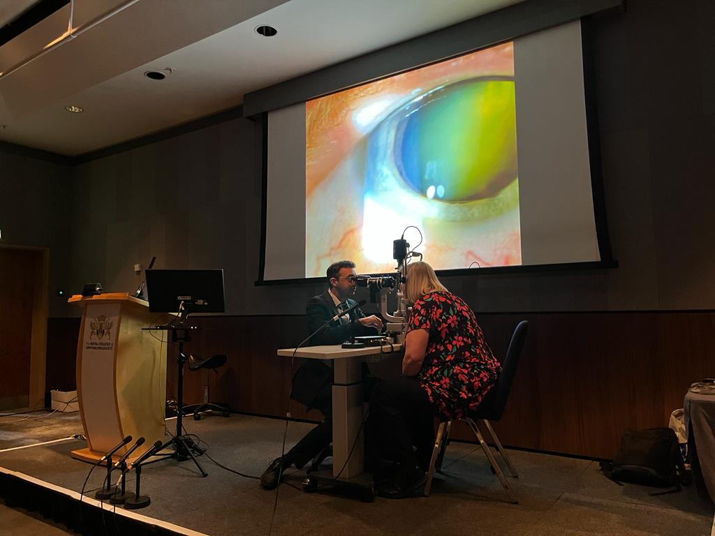 Black Country SLC member Clare Williams sat on a chair on stage. She is opposite Chair of the Royal College of Ophthalmologists Training Group, Sunil Mamtora, who is examining her eye. An image of Clare’s eye in the eye exam is displayed on a large screen for other conference delegates.