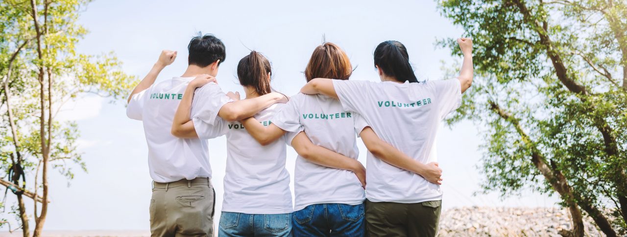 A photo of four people, taken from behind. They are wearing white t-shirts with the word 'volunteer' written across the back, in light blue. They are linking arms and the person on either end of the line has one arm raised, in a cheer.