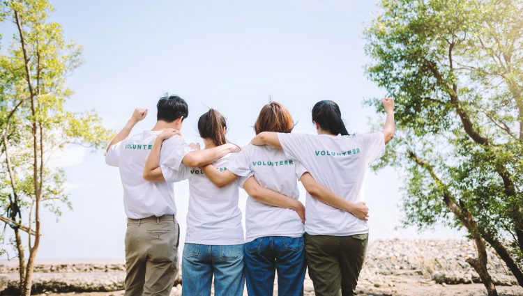A photo of four people, taken from behind. They are wearing white t-shirts with the word 'volunteer' written across the back, in light blue. They are linking arms and the person on either end of the line has one arm raised, in a cheer.