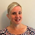 headshot of Kelly Barton, Engagement Manager for the north west. Kelly is standing against a white wall and smiling at the camera. Her hair is pulled back into a pony tail. She is wearing a black top with red and white flowers on it.