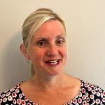 headshot of Kelly Barton, Engagement Manager for the north west. Kelly is standing against a white wall and smiling at the camera. Her hair is pulled back into a pony tail. She is wearing a black top with red and white flowers on it.