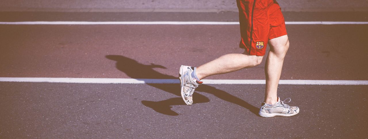 Image shows a man running on an athletics track from the waist down. He is wearing red shorts and white trainers.