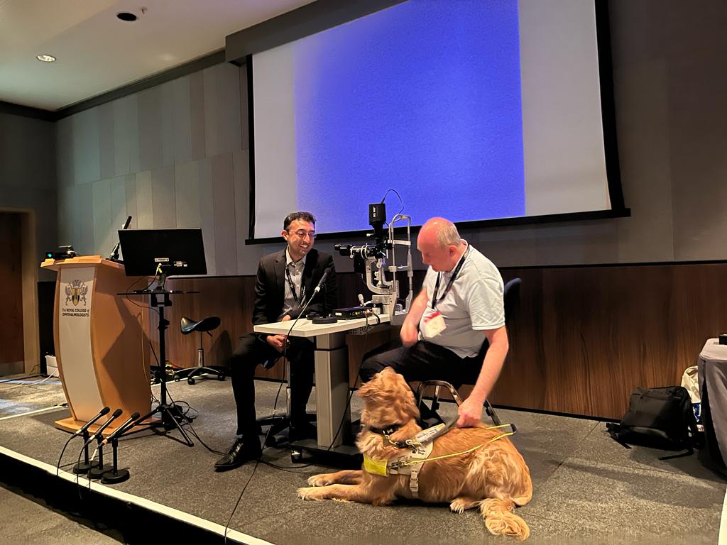 Birmingham SLC member Paul Hopkins sits on a chair on stage opposite Chair of the Royal College of Ophthalmologists Training Group, Sunil Mamtora, who is examining his eye. An image of Clare’s eye in the eye exam is displayed on a large screen for other conference delegates.