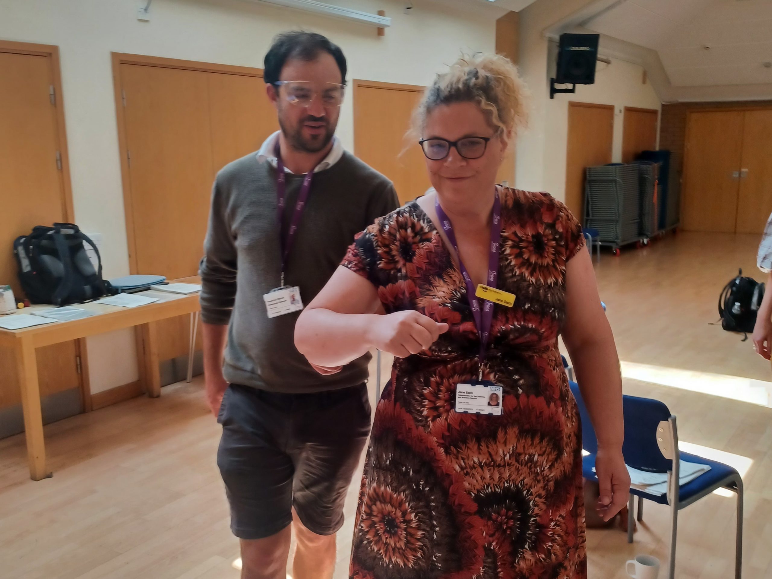 Two members of the Sirona DANS team are taking part in sighted guide training. The man is wearing sim specs, and being guided by a colleague.