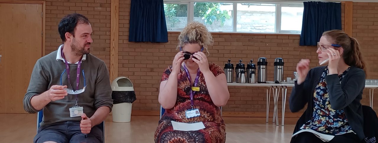 Three members from the Sirona Diabetes and Nutrition Services team are sat down, trying on simulation specs. They are talking to each other as they try them on.