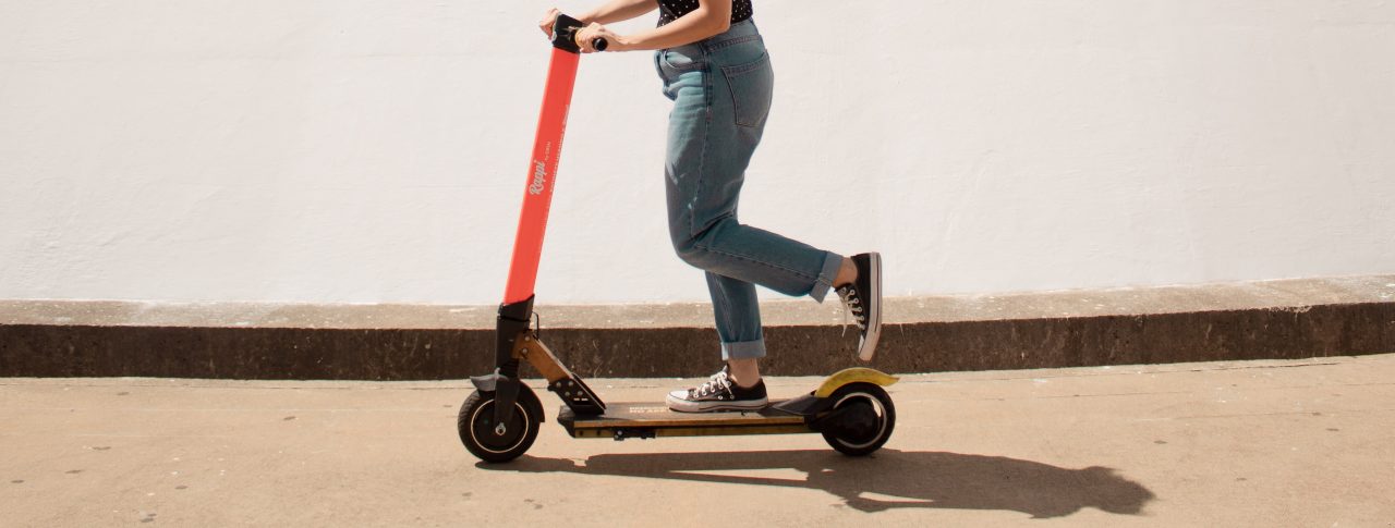 A lady is riding a red e-scooter on a path, against a white wall.