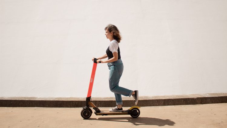 A lady is riding a red e-scooter on a path, against a white wall.