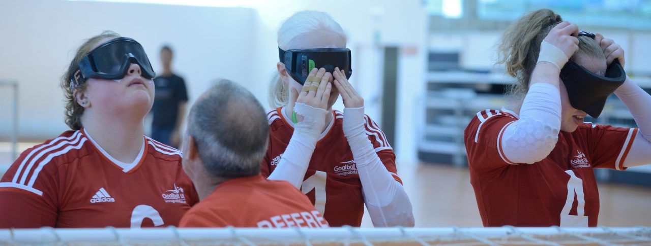 Three female Goalball UK players, standing behind a net. They are adjusting their blindfolds.