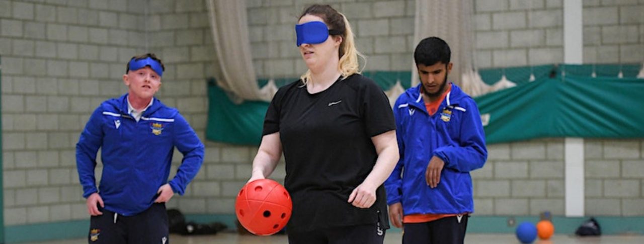 Three people playing Goalball. A lady at the front is wearing a blue blindfold and is holding a red ball. Two male team mates are standing behind her, in blue Goalball UK jackets.