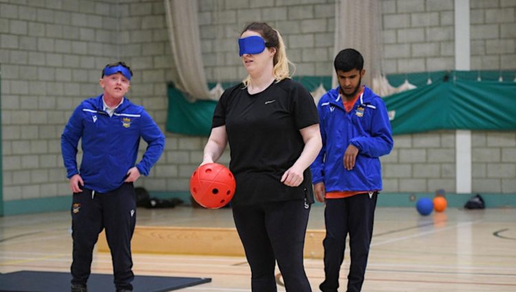 Three people playing Goalball. A lady at the front is wearing a blue blindfold and is holding a red ball. Two male team mates are standing behind her, in blue Goalball UK jackets.