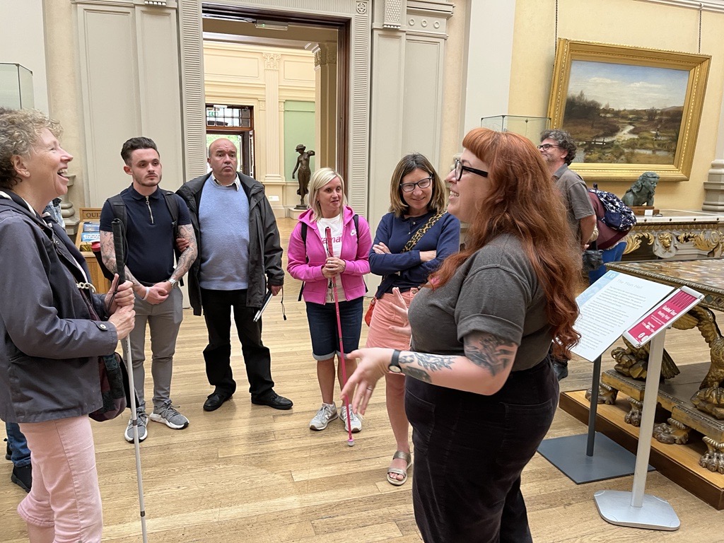 Members of Merseyside SLC at the Lady Lever Art Gallery. They are stood in a gallery, around a staff member, who is laughing with an SLC member during a conversation.