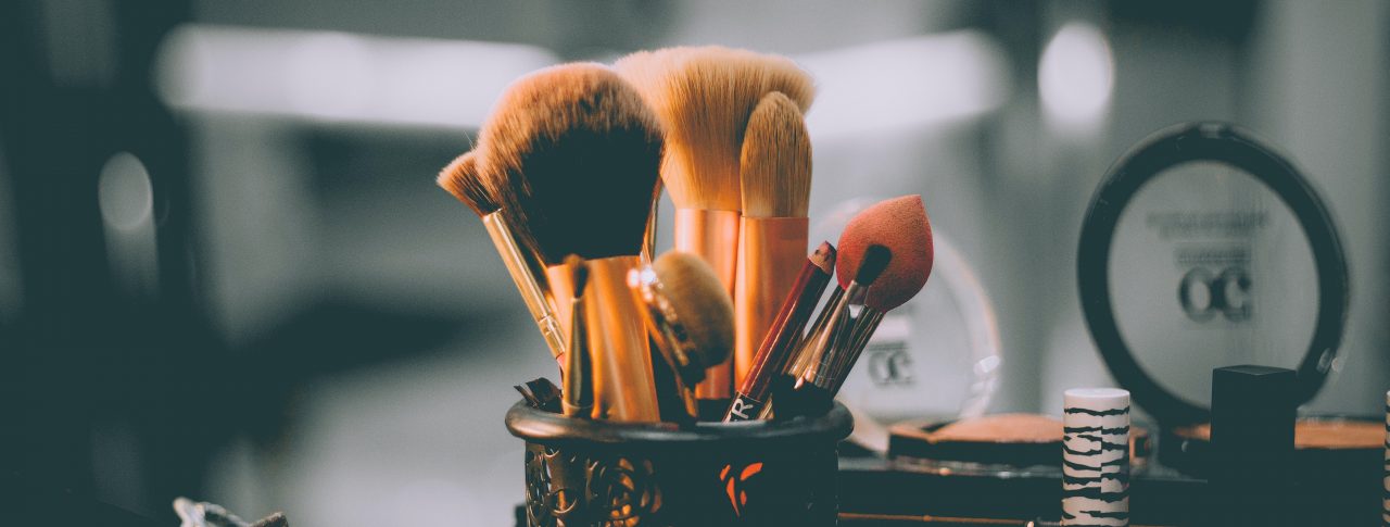 A selection of makeup brushes in a container on a desk. Other items of make up are shown in the background, highlighted by beauty mirror lights.