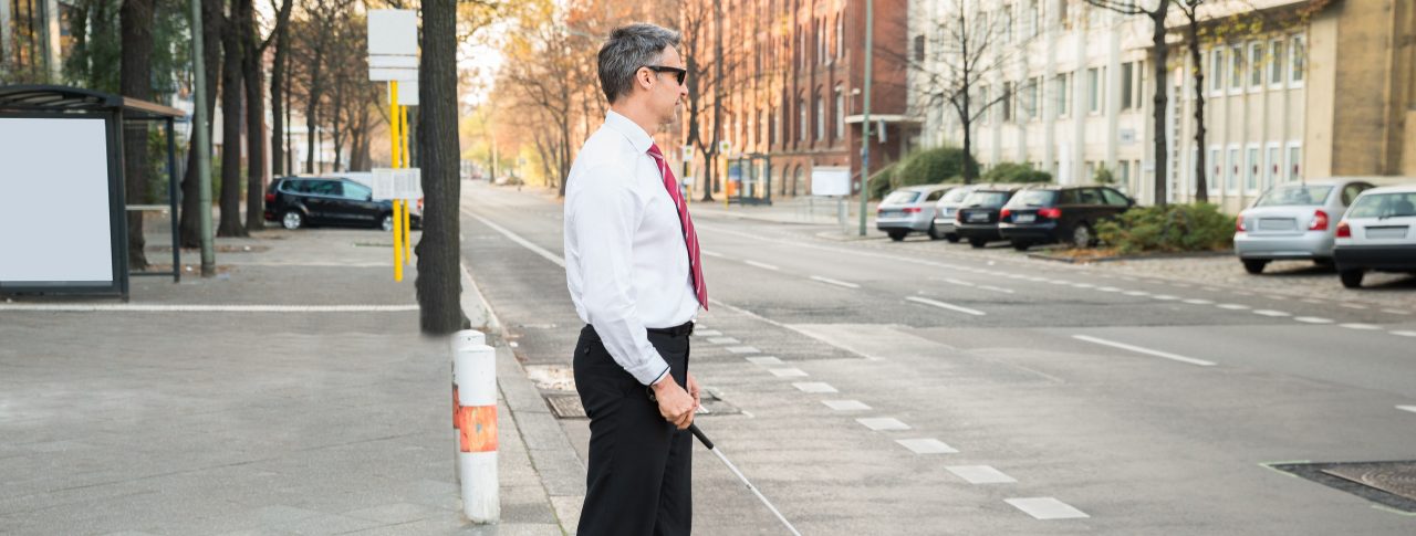 A man in a smart trousers, white shirt, and tie is standing on the edge of a curb holding a cane. He is about to cross a road, via a bus lane. Trees, houses, and some cars line either side of the road.