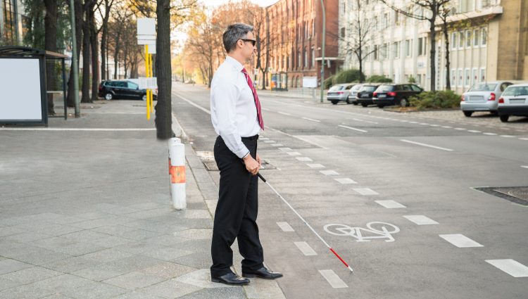 A man in a smart trousers, white shirt, and tie is standing on the edge of a curb holding a cane. He is about to cross a road, via a bus lane. Trees, houses, and some cars line either side of the road.