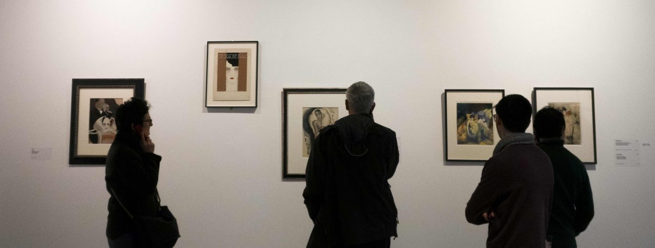 Four people looking towards five small pieces of art, in black frames, on a grey wall.