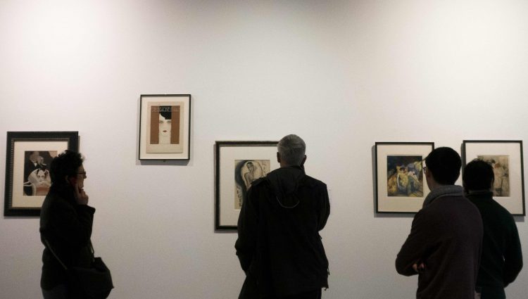 Four people looking towards five small pieces of art, in black frames, on a grey wall.