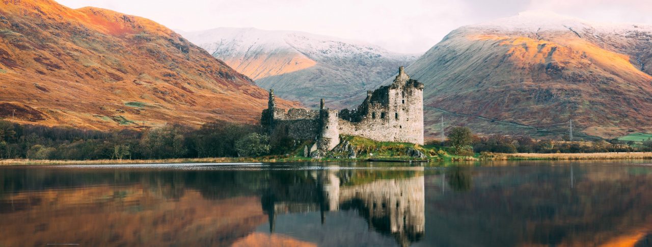 Image looks across a loch, at a castle nestled in front of three hills. The sun is setting, and the reflection of the scene can be seen in the water.