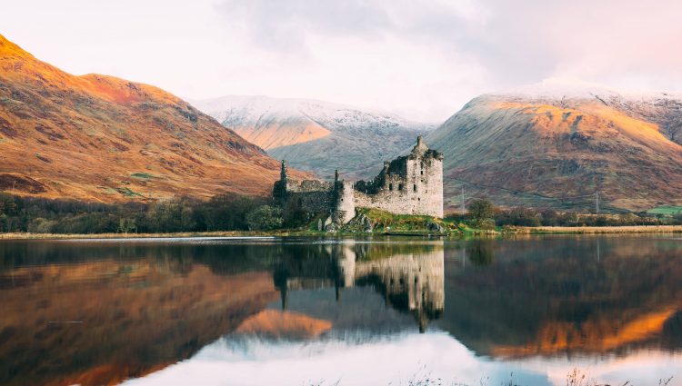 Image looks across a loch, at a castle nestled in front of three hills. The sun is setting, and the reflection of the scene can be seen in the water.