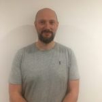 Headshot of Steven Bignell, Merseyside SLC member. He is standing against a cream wall, facing the camera, smiling. He is wearing a grey t-shirt.