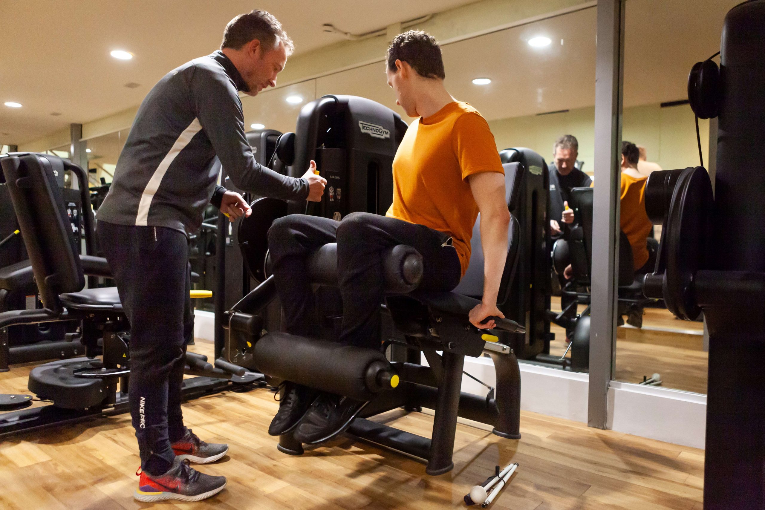A visually impaired male, sitting on a piece of gym equipment. An instructor is standing with him, showing him how to use the equipment.
