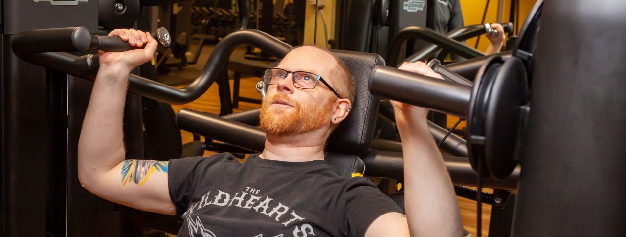 A partially sighted male, using weights in the gym. An instructor is seen in the background, smiling encouragingly.
