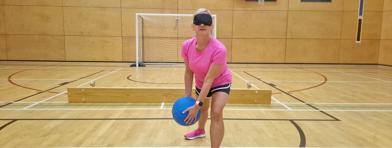 Kelly Barton, Engagement Manager for the North West is stood poised holding a goalball, on the court. She is wearing a bright pink t-shirt and a black blindfold.