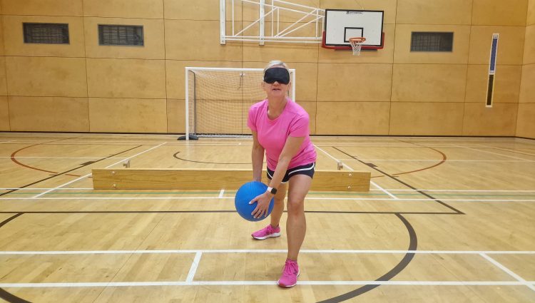 Kelly Barton, Engagement Manager for the North West is stood poised holding a goalball, on the court. She is wearing a bright pink t-shirt and a black blindfold.