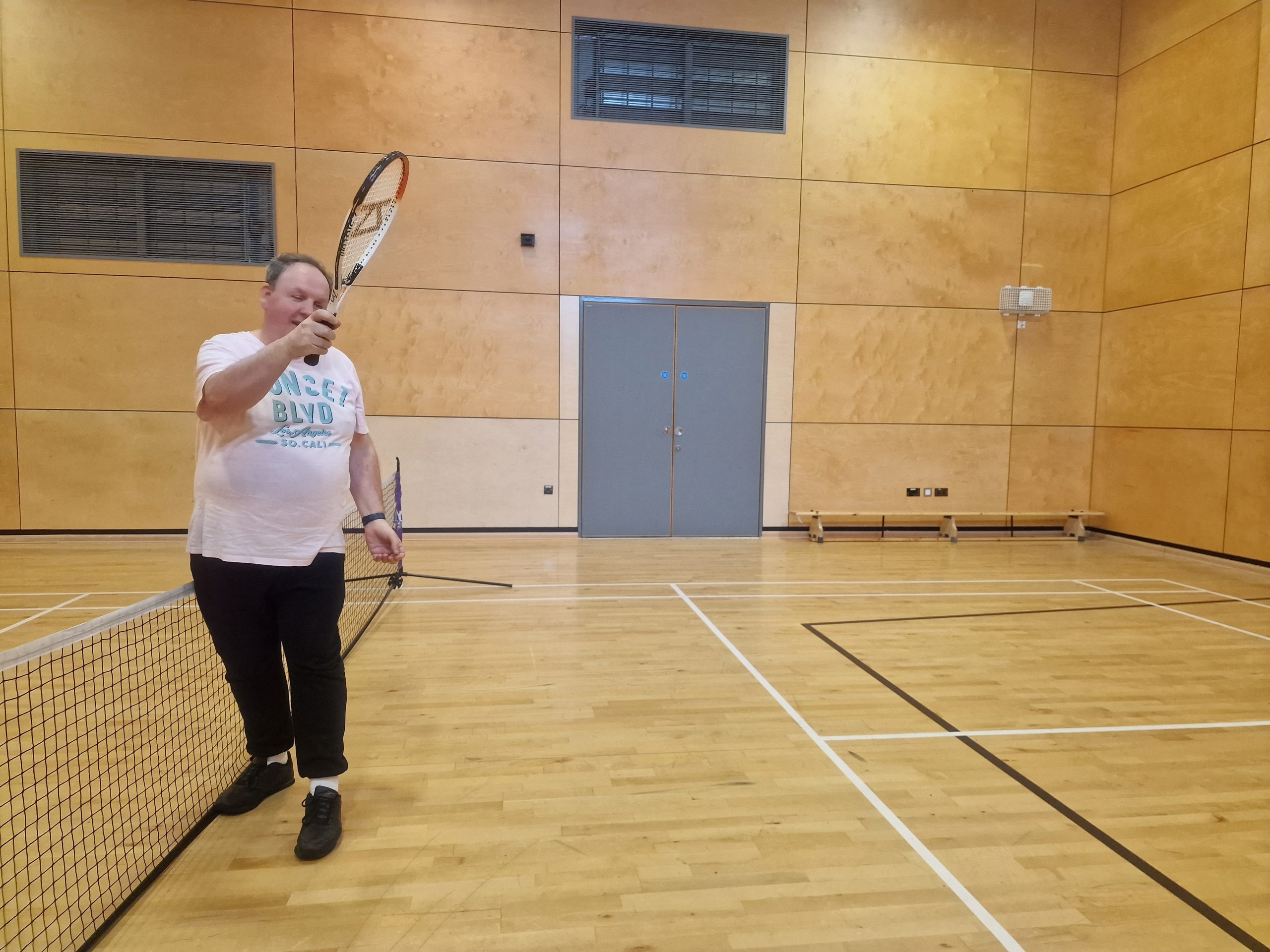 Participant Michael, is shown standing next to the net, swinging his tennis racket up, during the VI tennis session.