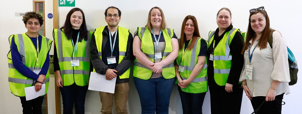 Six male and female sighted guides stood in a row smiling at the camera. They are pictured with Sight Loss Council Engagement Manager Sam, pictured with her guide dog.