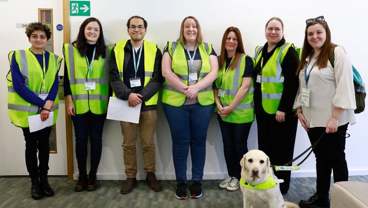 Six male and female sighted guides stood in a row smiling at the camera. They are pictured with Sight Loss Council Engagement Manager Sam, pictured with her guide dog.