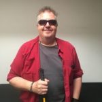 Photo of James Connell, Merseyside SLC member. James is standing against a wall, looking at the camera. He is wearing sunglasses and holding his cane in one hand. James is smiling at the camera.