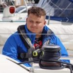 Photo of Lloyd Sakr, Lancashire SLC member. Lloyd is sitting behind ropes on a sail boat, wearing a blue jacket.