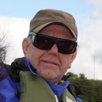 Photo of Micahel Parkinson, Lancashire SLC member. Michael is outside, wearing a khaki life jacket over a blue coat. He is wearing a khaki cap and black sunglasses.