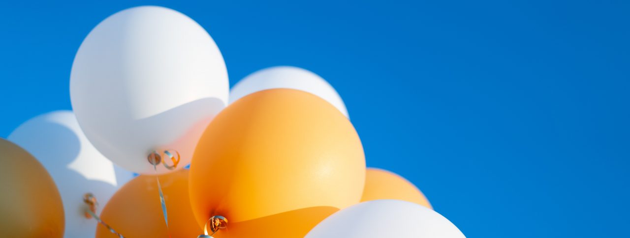 A bunch of white and yellow balloons, tied together with gold string, held up against bright blue sky.