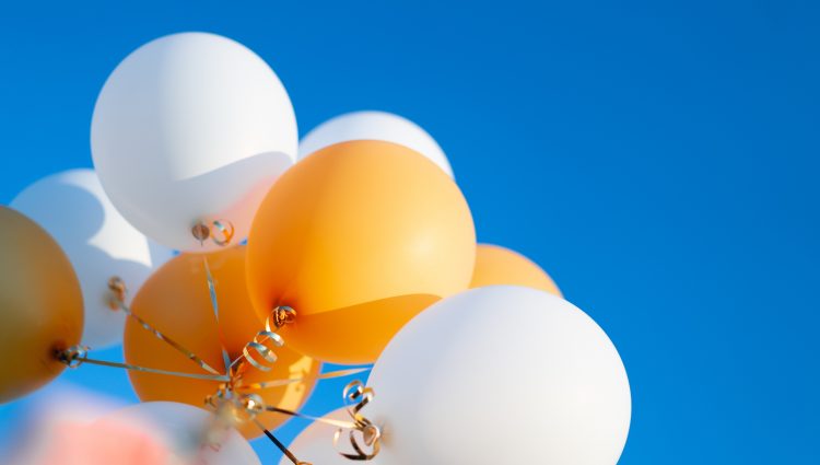 A bunch of white and yellow balloons, tied together with gold string, held up against bright blue sky.
