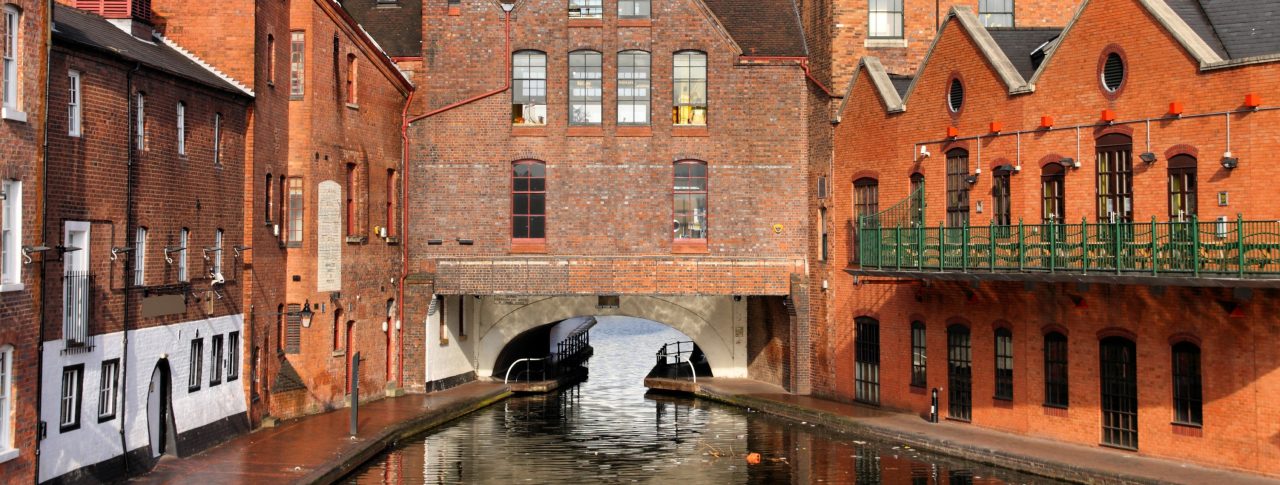 Image of a canal in Birmingham. Old, red brick buildings line the pathways either side, and there is an archway under an old house in the centre of the image.