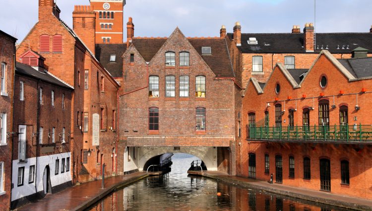 Image of a canal in Birmingham. Old, red brick buildings line the pathways either side, and there is an archway under an old house in the centre of the image.