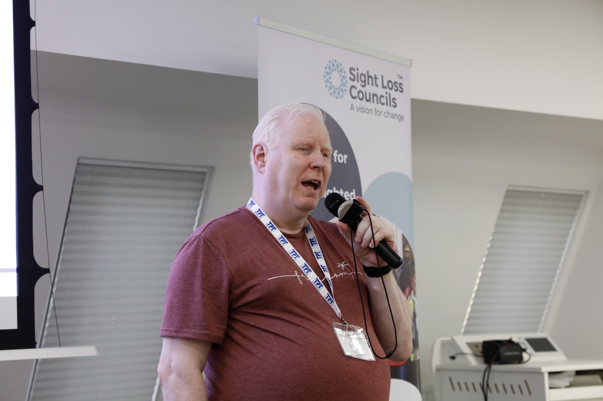 Iain Mitchell, Senior Engagement Manager for the North, speaking into a microphone. A SLC banner is behind him.