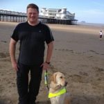 Image shows James Preston with his guide dog on a sandy beach. There is a pier in the background.